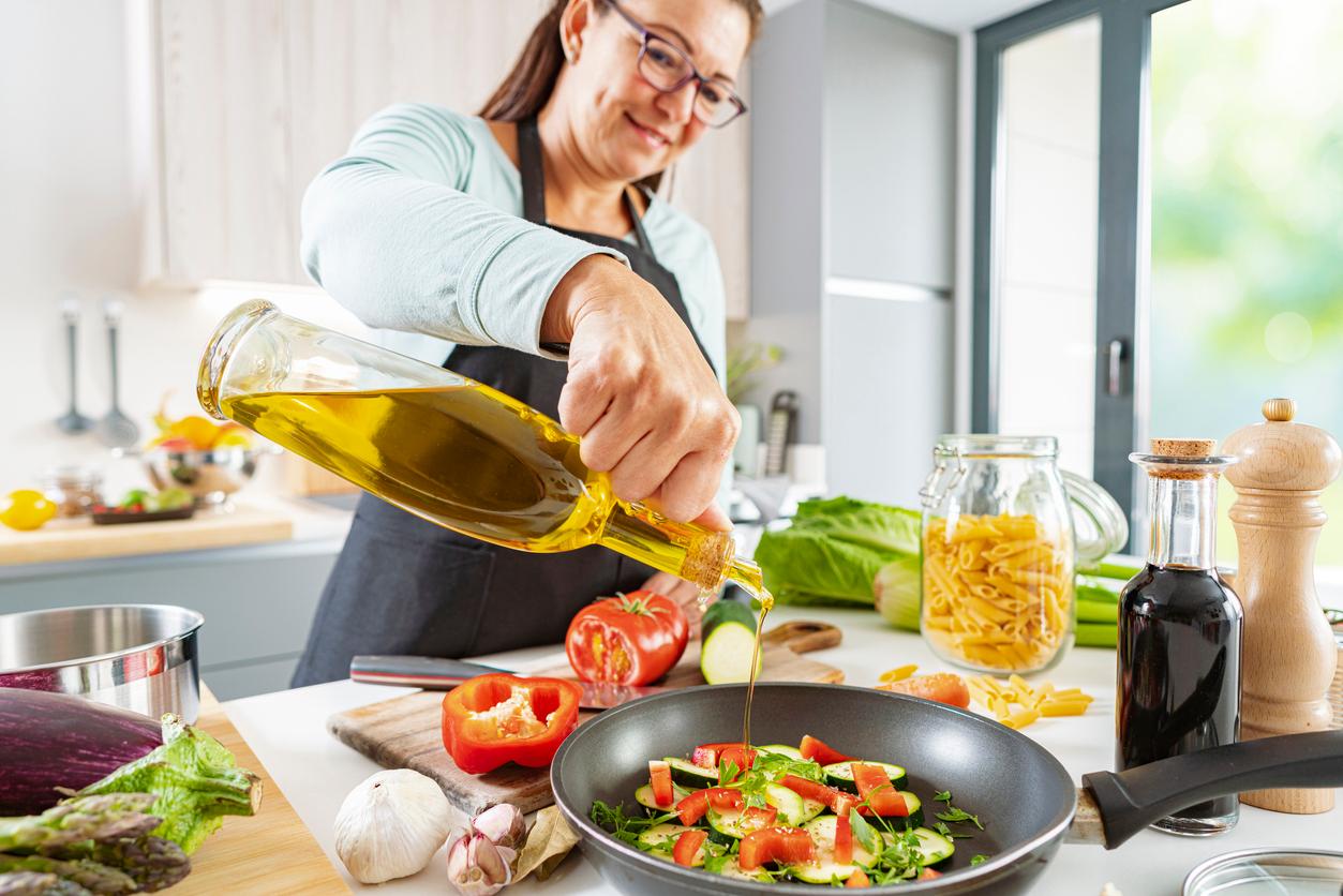 A chef inside a kitchen pours olive oil into a pan of veggies atop a counter that has tomatoes and other vegetables as well as a jar of pasta.